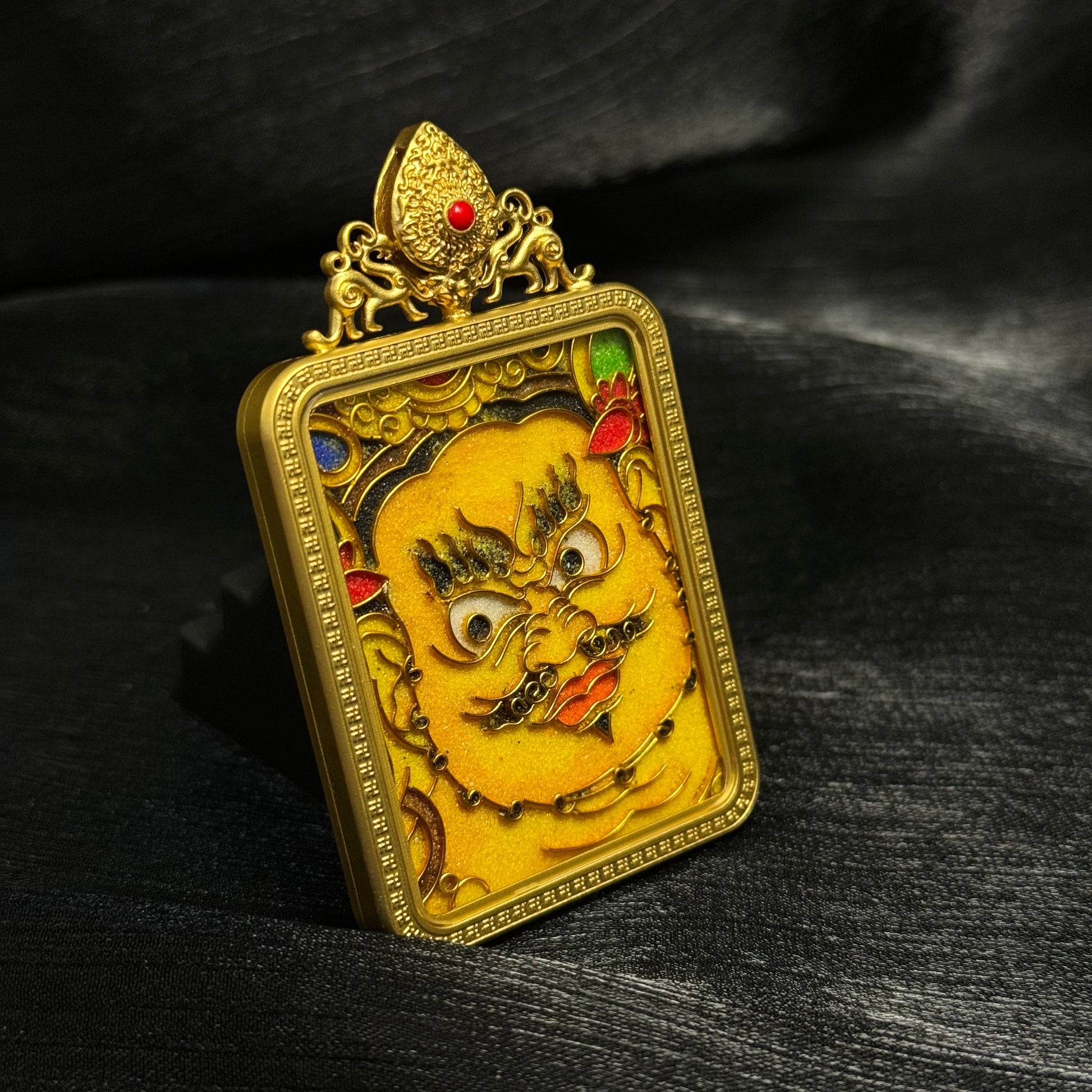 Close angled perspective of Yellow Jambhala cloisonné enamel pendant, showing textured enamel surface, gold-plated frame, and symbolic wealth deity imagery.